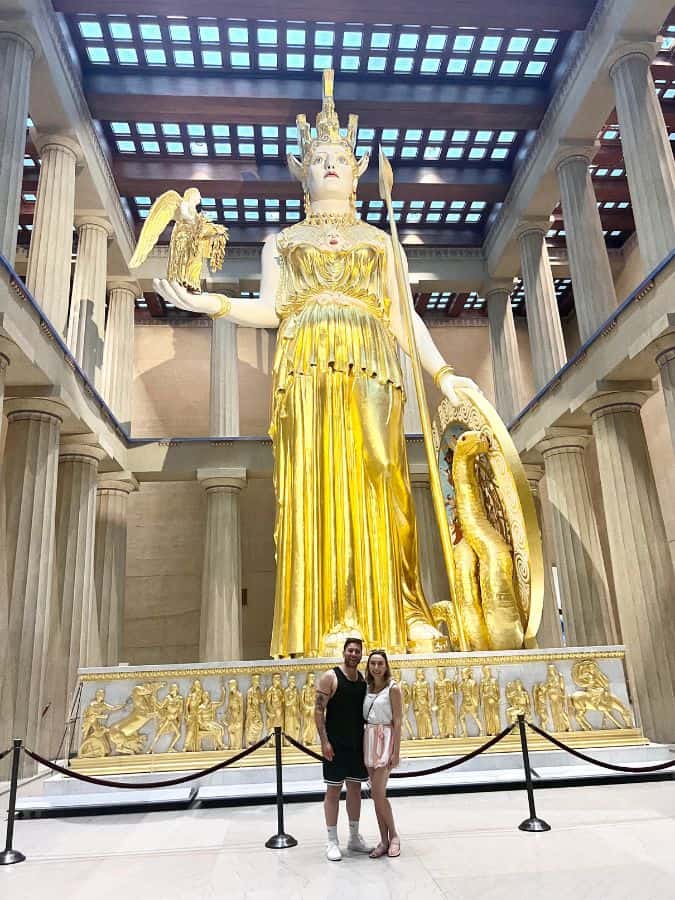 photo of the athena statue inside the parthenon nashville tennessee. It is a huge statue and covered in gold. There is a couple standing in front of the statue smiling. 
