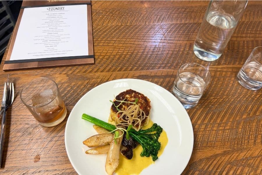 A dinner plate with a crab cake, trumpet mushrooms, broccolini, and a yellow sauce plated on it. There is a glass with bourbon to the side as well as a water glass. In the corner is the food menu