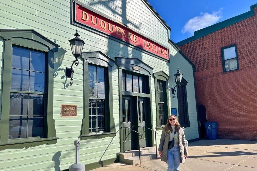 A young woman outside in front of a green building that reads "Duquesne Incline". This is the historical building at the top of Mount Washington in Pittsburgh PA