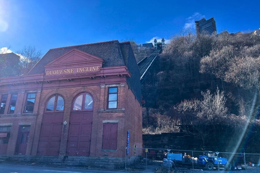 The outside of the Duquesne Incline building in Pittsburgh Pennsylvania. The building is red brick and it says "Duquesne Incline" on it. Behind is a track that conveys the cart up a hill to Mount Washington