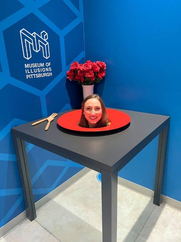 A girl with her head coming through a table as an optical illusion. On the wall behind her is a sign that reads "Museum of Illusions"