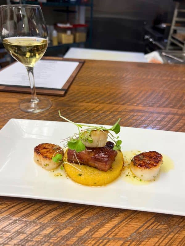 Scallop dish with polenta and bbq pork belly. Plated professionally in a restaurant paired with a glass of white wine