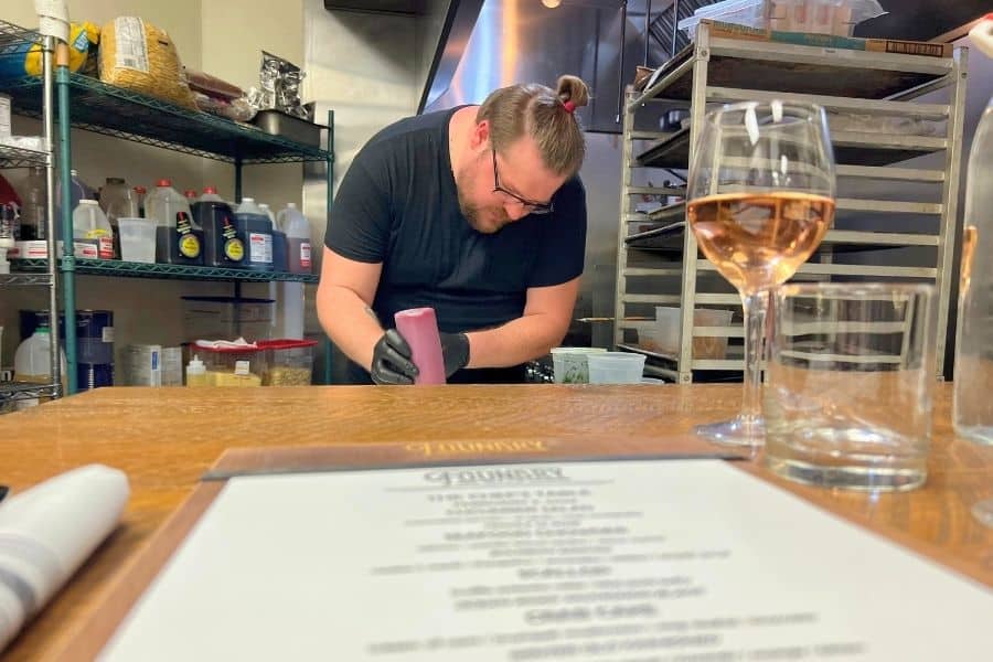 A food menu and a glass of wine with a chef preparing a dish behind it. This is a photo of a private chefs table experience held in a restaurant kitchen