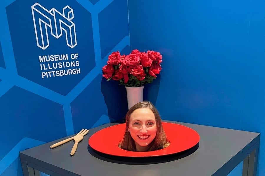 A girl with her head poking up inside a plate to make it look like she is on the dinner plate. The sign "Museum of Illusions Pittsburgh" is behind her